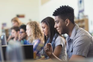 Young adult or teenage African American male college student is smiling while using a computer in college computer lab. Young man is sitting at desk in a row with diverse classmates. They are wearing casual clothing and looking at computer screens.